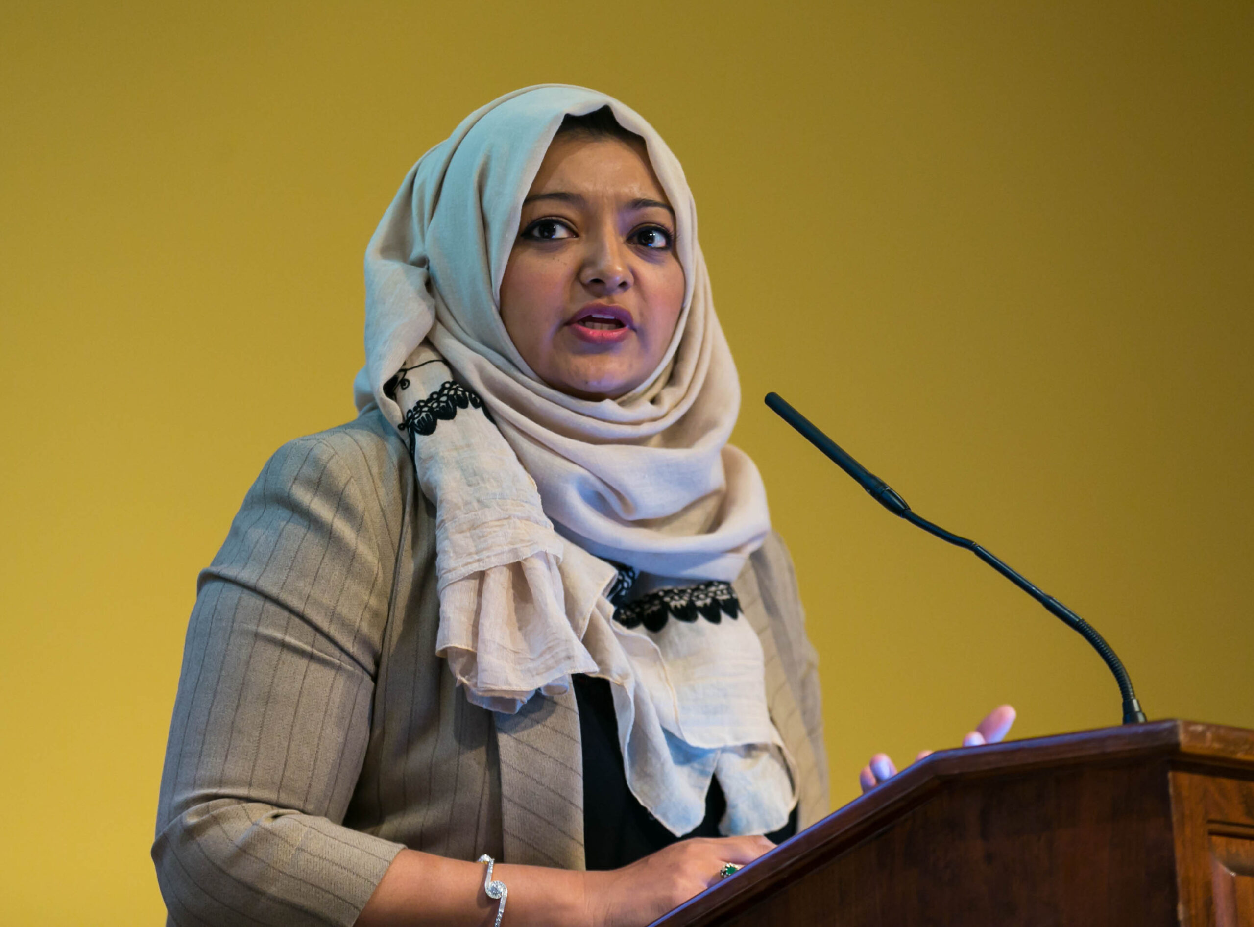 Rabia Chaudry wearing a beight short and head scarf speaks into a microphone.